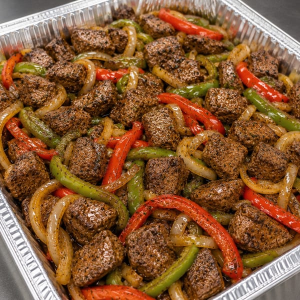 Close-up of steak bites with red and green bell peppers and onions in a foil pan.