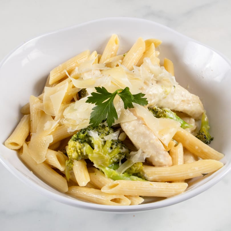 Penne pasta with chicken, broccoli, creamy sauce, and shaved Parmesan cheese in a white bowl.